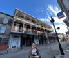A person stands in a sunny street with traditional wrought-iron balconies in what appears to be the French Quarter of New Orleans
