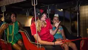 A group of individuals appears to be on a bus or tram ride at night, with a person in a red shirt and black hat seemingly acting as a tour guide or entertainer.