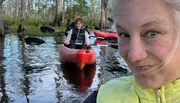 A woman is taking a selfie in the foreground while another person paddles a kayak in a swampy waterway behind her.