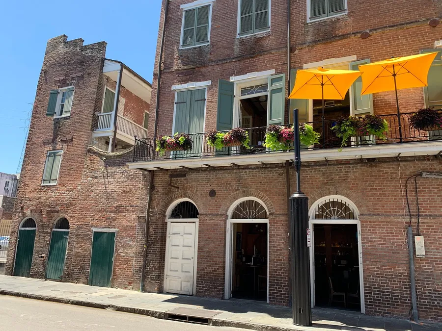 The image shows a sunny street view of a charming brick building with French doors, balcony planters filled with flowers, and bright yellow umbrellas, suggesting a quaint urban area, possibly in a historic district.