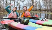 Two happy people are kayaking together in a colorful tandem kayak, waving cheerfully in a scenic wetland environment.