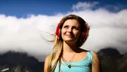 A young woman is wearing red headphones and smiling while looking up towards the sky with clouds and mountains in the background.