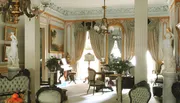 The image displays an opulent room with classical decor, featuring fluted columns, ornate furniture, statues, and drapery, evoking the style of a bygone era.