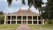 This image shows a symmetrical two-story building with a red-tiled roof, large double-decker porches lined with columns, and Spanish moss hanging from trees in the foreground.