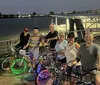 A group of six people posing with bicycles adorned with colorful LED lights by a riverfront at dusk
