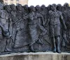 The image shows a bronze bas-relief sculpture depicting a group of expressive interconnected figures that appear to reflect a historical or cultural narrative