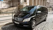 A black Mercedes-Benz Viano van is parked on a cobblestone street next to a sidewalk with metal railings, and a historic building can be seen in the background.