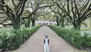 A person stands on a brick pathway lined with large, gnarled oak trees leading towards a classic white building with a veranda.