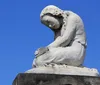The image shows a weathered white stone sculpture of an anguished figure hunched over in a pose that suggests deep sorrow set against a clear blue sky