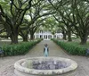 The image shows a pathway lined with majestic trees leading to an elegant white house with a small figure standing near a stone fountain in the foreground