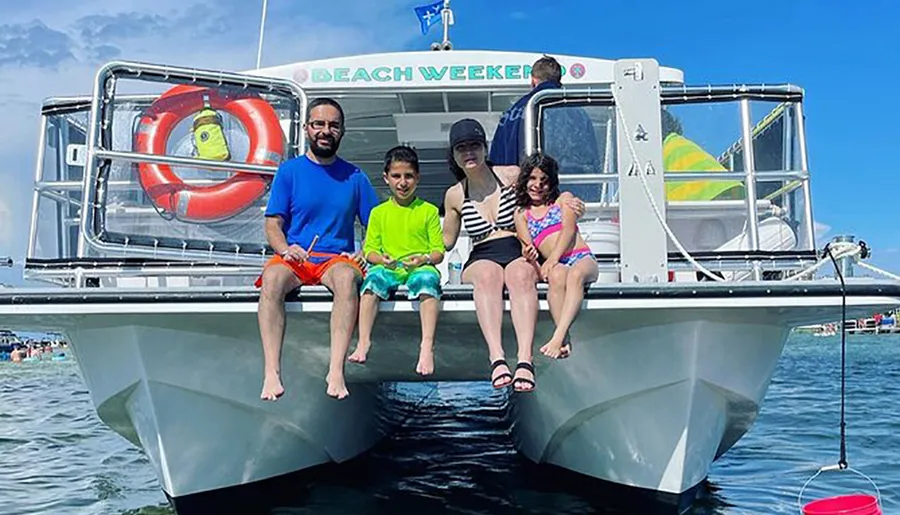 A family is sitting at the bow of a catamaran style boat named BEACH WEEKER, smiling and posing for a photo on a sunny day.
