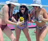 Three women in swimsuits and sun hats celebrate with a drink on a sunny beach day