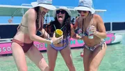 Three women in swimsuits and sun hats celebrate with a drink on a sunny beach day.