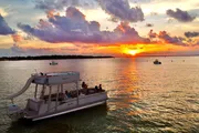 A pontoon boat with passengers cruises on a calm waterway against the backdrop of a vivid sunset and scattered clouds.