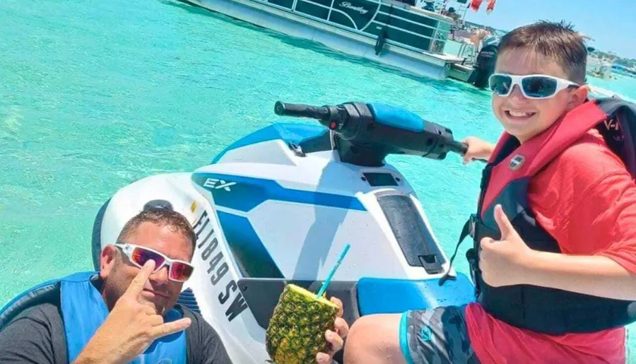 A man and a young boy are posing with thumbs up next to a jet ski in clear blue water, with the man holding a pineapple drink.