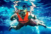 A person is submerged in clear blue water, giving two thumbs up while wearing snorkeling gear and an orange life vest.