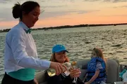 A server is pouring a beverage for a smiling woman on a boat during a sunset cruise, with a child seated beside her looking on.