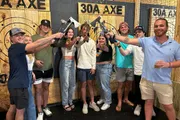 A group of smiling people are posing for a photo while holding axes at an indoor axe-throwing venue.