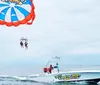 Two people are parasailing above the ocean towed by a boat that is moving swiftly across the water
