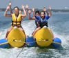 Three children are joyfully riding a banana boat on the water with their arms raised in excitement