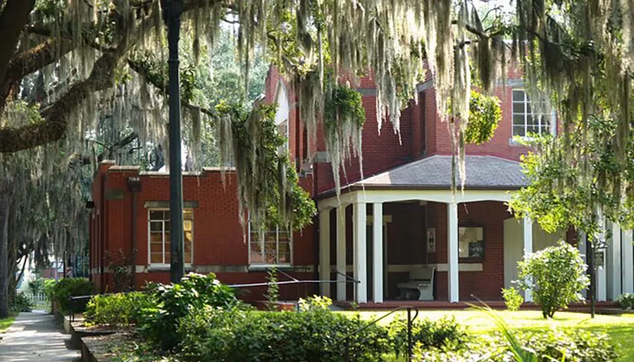 A red brick building with white columns and a porch, shaded by Spanish moss-draped trees, exudes a tranquil, Southern charm.