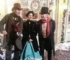 Three individuals are dressed in historical costumes exuding a festive spirit with one playing a violin amidst a decorated snowy setting reminiscent of a Charles Dickens novel