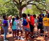 A group of diverse students on a sunny day is engaged in an outdoor educational activity led by a guide gesturing expressively