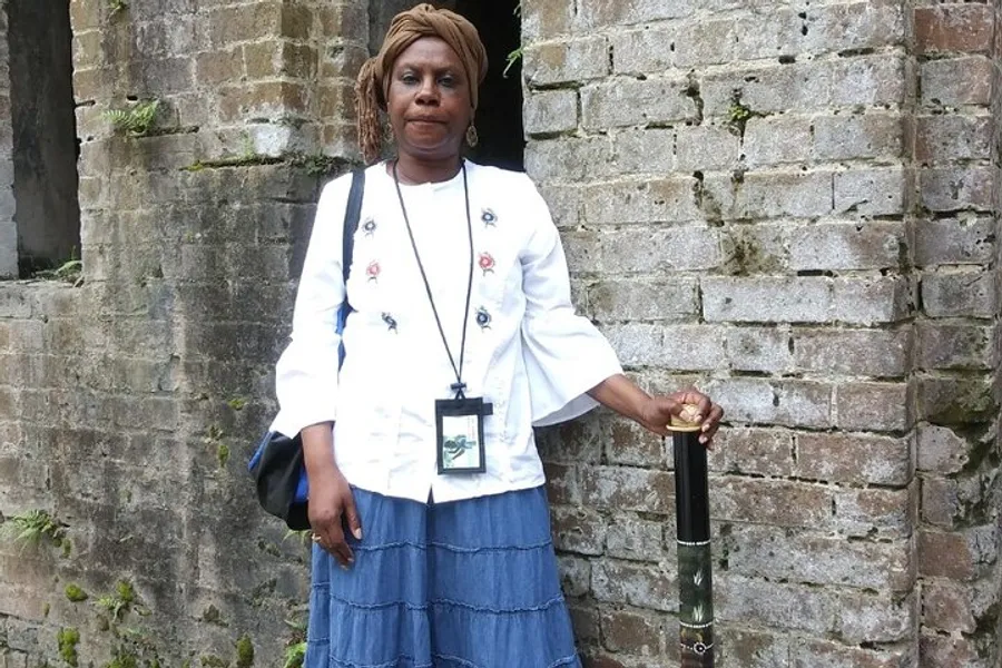 A woman is standing against a brick wall with her hand resting on an umbrella, wearing a white blouse with floral detail, a blue skirt, and a badge around her neck.