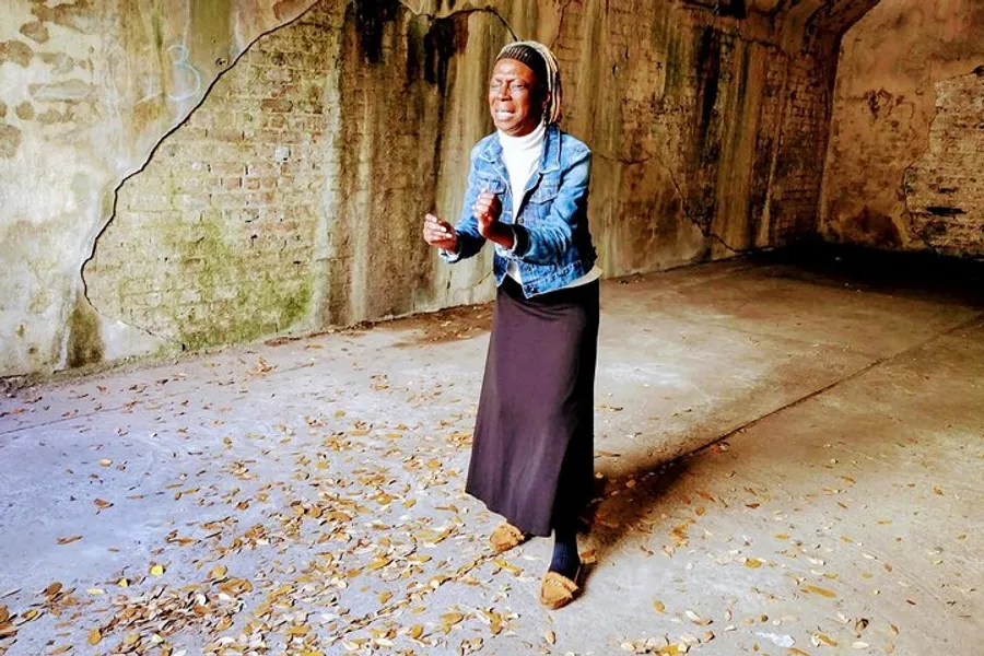 A person is standing on a floor covered with fallen leaves inside a room with weathered walls, holding an object and appearing to be in mid-speech or singing.