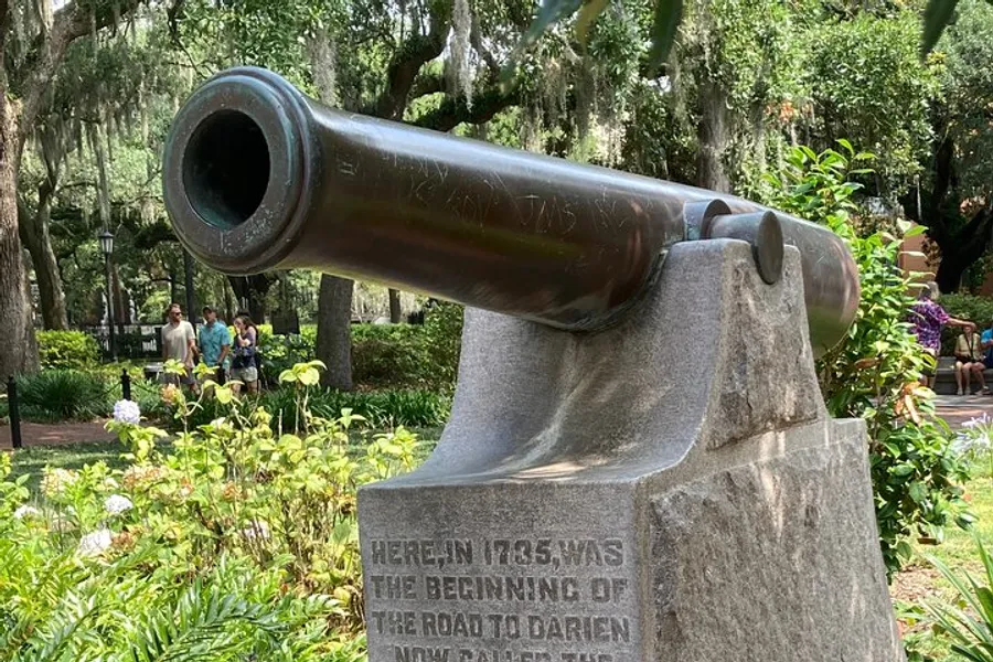 The image shows an old cannon mounted on a stone pedestal with an inscription, set in a lush park with people in the background.