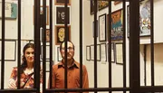 A man and a woman stand behind a set of vertical bars, with various framed artworks on the wall behind them.