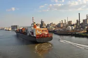 A large cargo ship loaded with containers is navigating a river near industrial facilities while a small boat speeds by in the foreground.