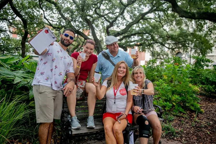 Five cheerful adults are posing for a photo on a park bench, holding drinks and a clipboard, surrounded by greenery.