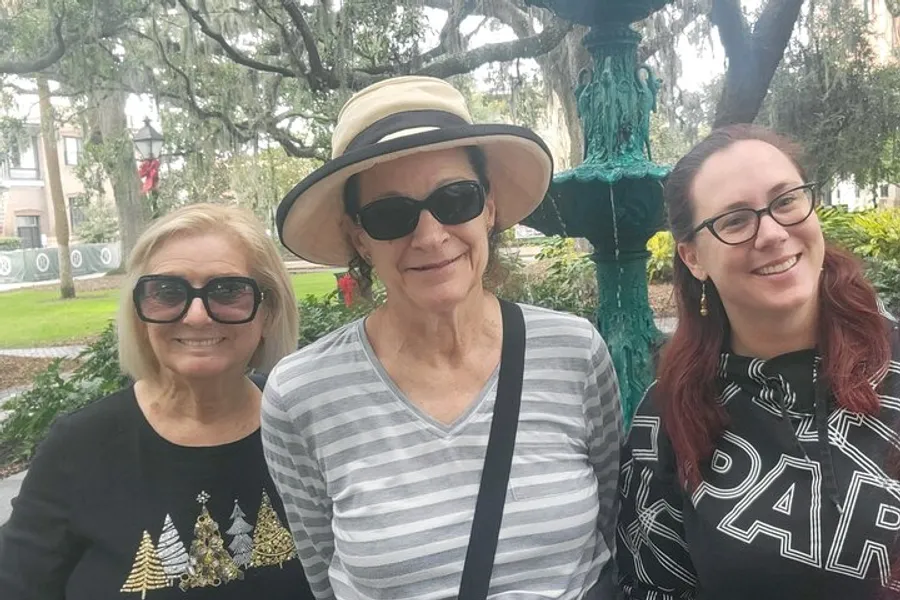 Three smiling women pose together for a photo in an outdoor setting with trees and a decorative lamp post in the background.