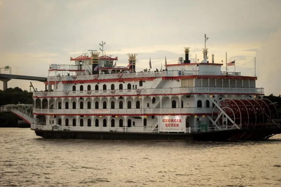 The image displays the Georgia Queen, a large paddlewheel riverboat with multiple decks, cruising on a river against an overcast sky.