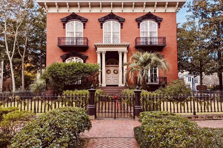 A stately red brick house with ornate black wrought-iron details and a well-manicured garden stands under a clear sky.