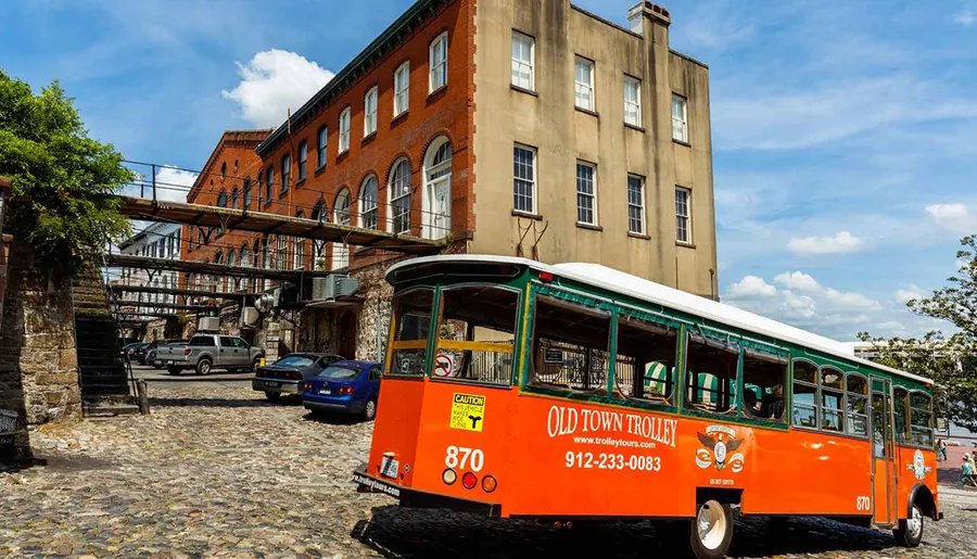 An orange and green old-fashioned trolley bus marked Old Town Trolley tours through a cobblestone street beside historical buildings under a sunny sky.