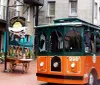 An orange and green trolley bus is parked on a cobblestone street next to a pedestrian and various storefronts