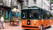 An orange and green trolley bus is parked on a cobblestone street next to a pedestrian and various storefronts.