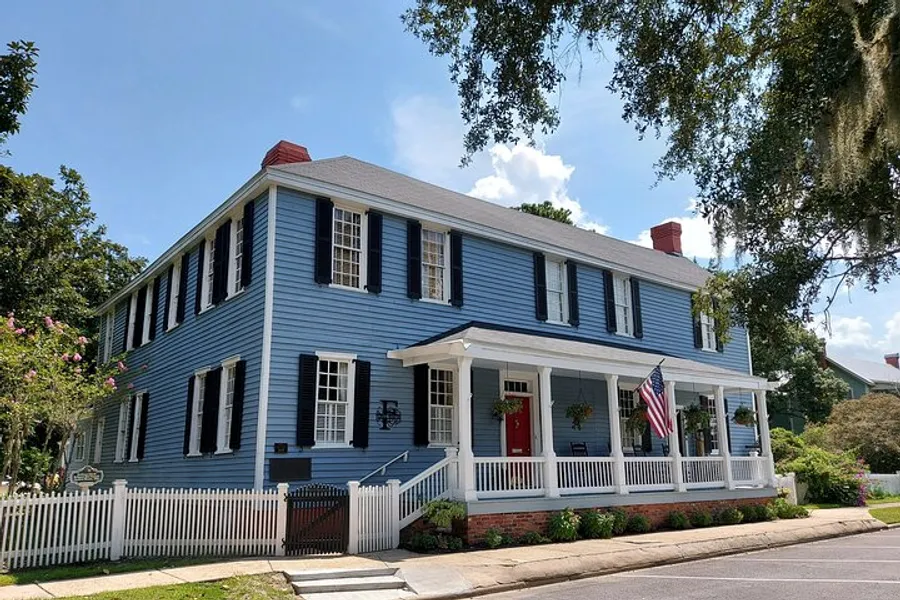 A classic two-story blue house with white trim, a red front door, an American flag, and a welcoming front porch under a sunny sky.
