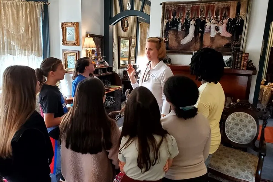 A group of children attentively listens to a woman who appears to be giving a tour or a presentation in a room adorned with antique furniture and framed artwork.