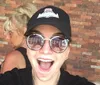 A person in sunglasses and a cap is smiling widely for a selfie with another person slightly out of focus in the background against a brick wall