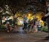 A group of people attentively listens to a person who appears to be telling a story or guiding a tour at night in a park illuminated by soft lights and draped with Spanish moss