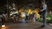 A group of people attentively listens to a person who appears to be telling a story or guiding a tour at night in a park illuminated by soft lights and draped with Spanish moss.