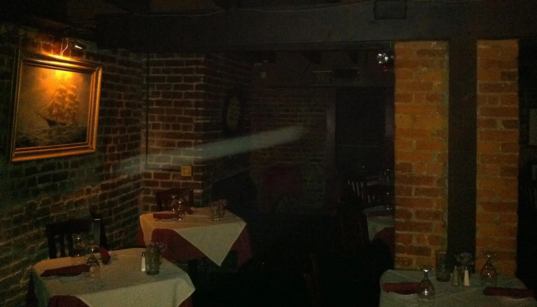 This image portrays a dimly lit, seemingly empty restaurant with brick walls, framed artwork, and tables set for dining.