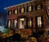 A stately two-story house with warm interior lighting is photographed at night showcasing its classic architecture and lush garden under a twilight sky