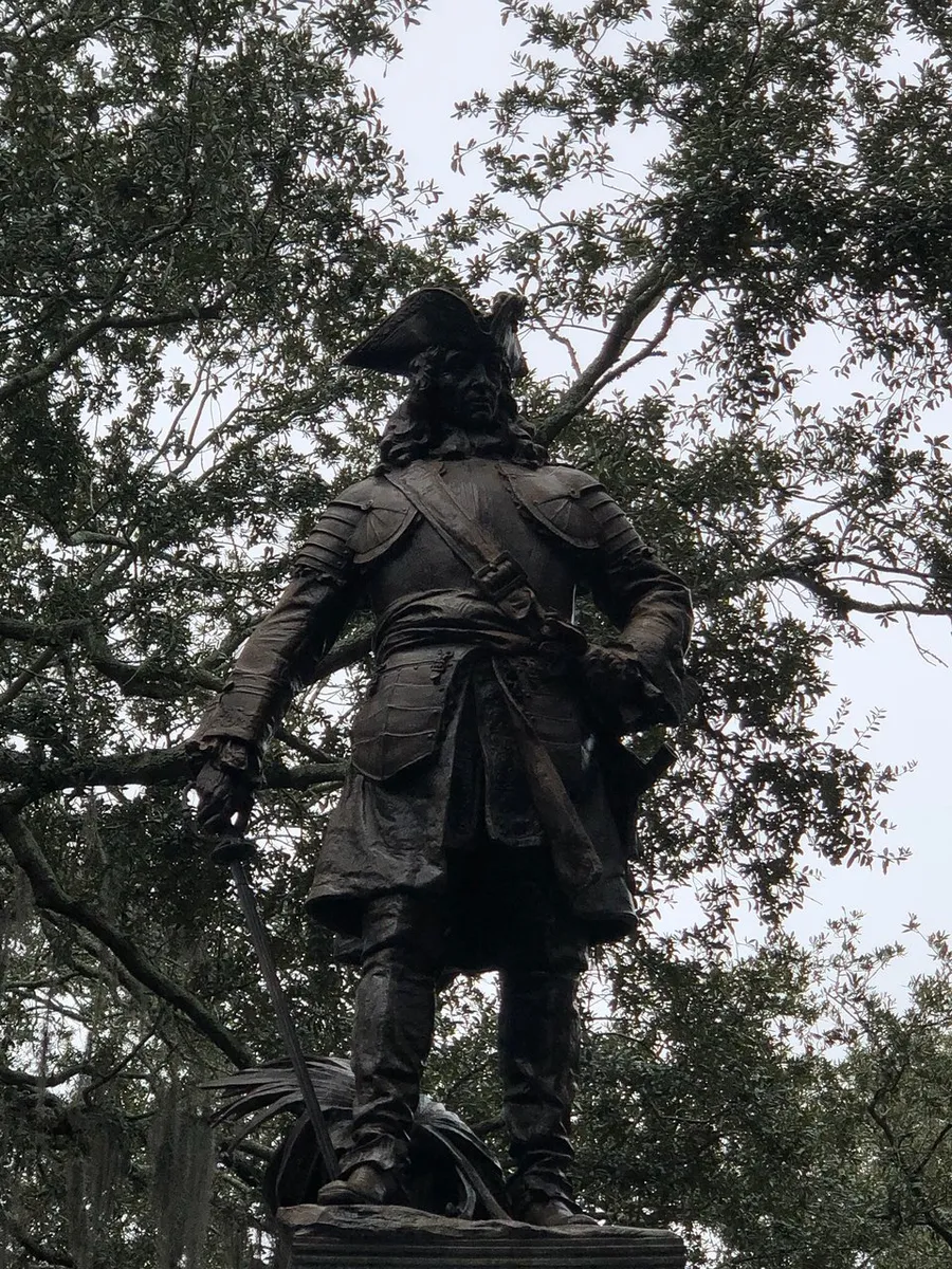 The image shows a statue of a historical figure in traditional attire, including a tricorne hat and a sword, set against a backdrop of leafy trees.