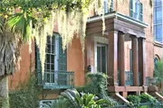 The image shows a charming, historic-looking terracotta-colored house with green shutters, ornate ironwork on the balcony, surrounded by lush greenery and draped with Spanish moss.