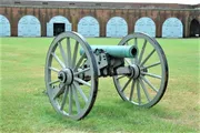 An old-fashioned cannon on wooden wheels is displayed on the grass in front of a building with rounded, blue doors.