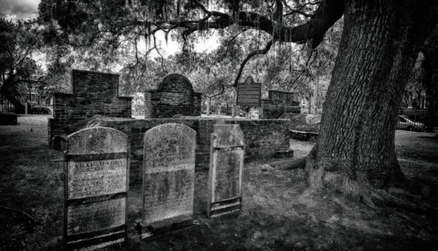 The image shows a serene, black and white scene of an old graveyard with weathered tombstones, overgrown trees, and aged brick structures giving it a somber and historical ambiance.